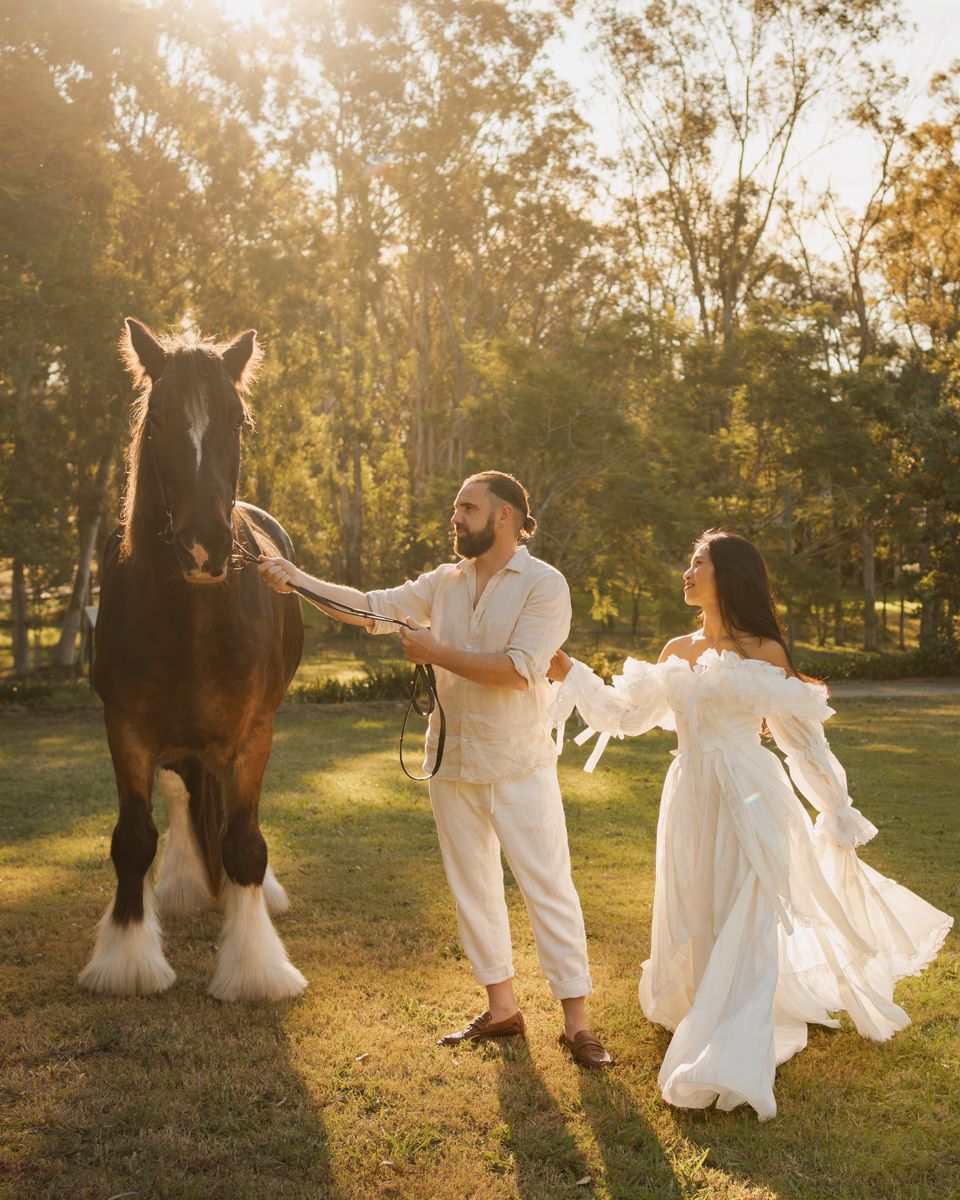 Couple in white attire posing with a brown shire horse at golden hour in a lush green park during a photoshoot with a horse in sydney