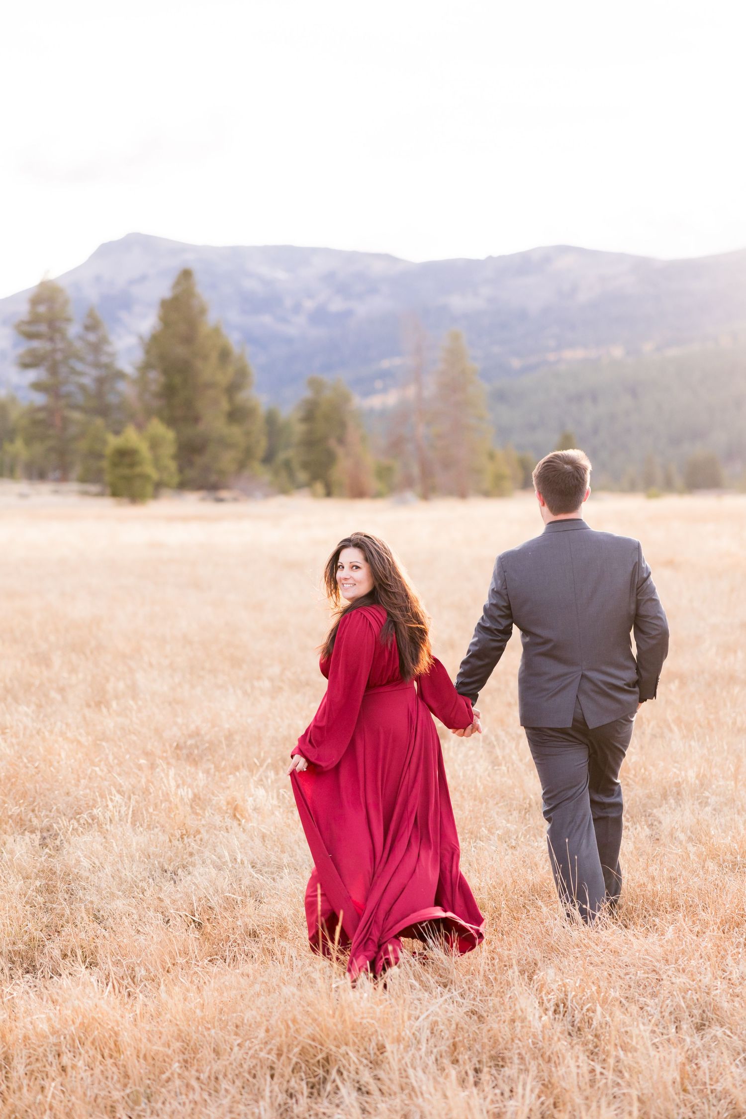 Couple walking hand-in-hand through golden meadow, woman in flowing red gown glancing back, mountains and pines behind.