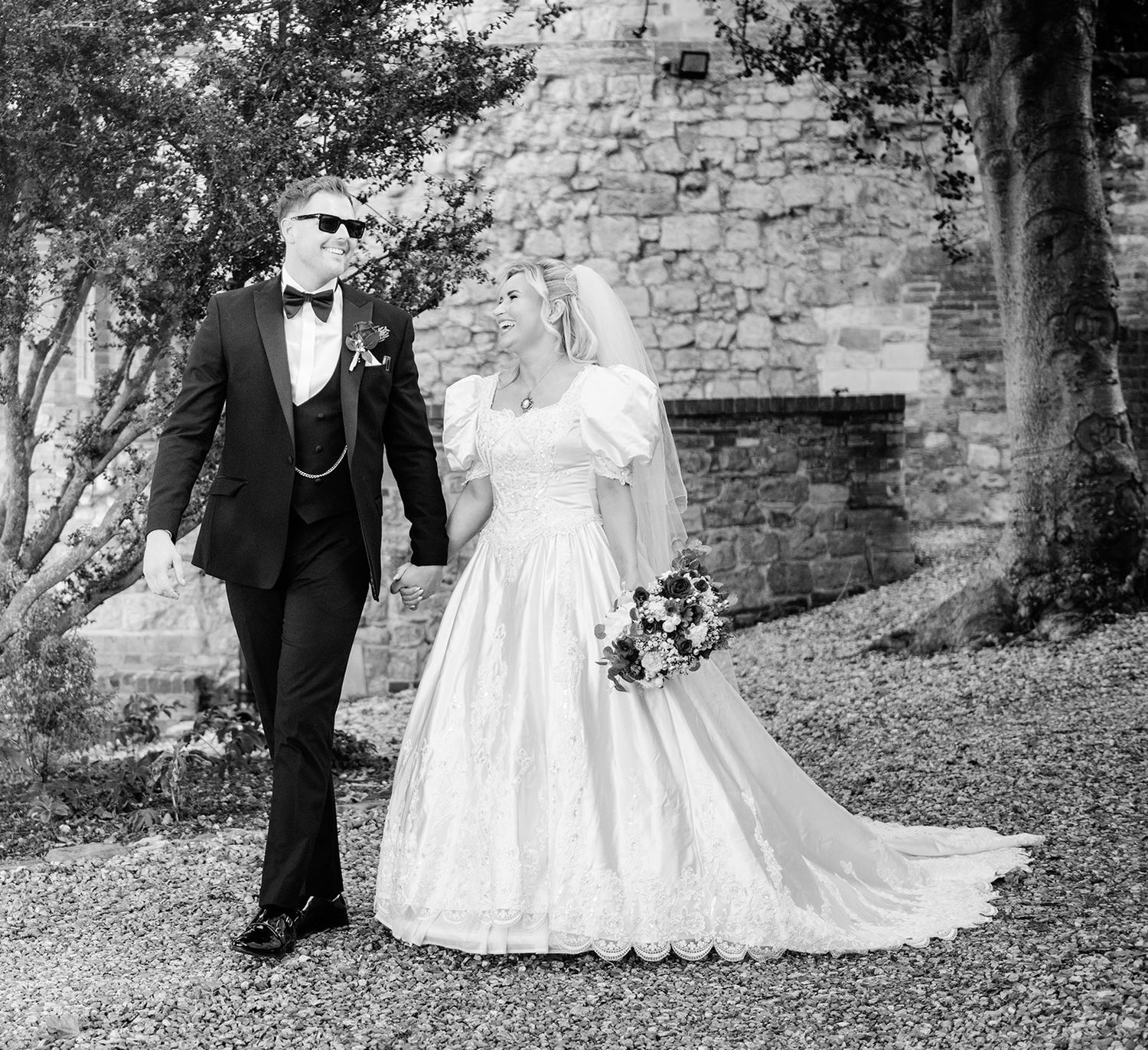 Black and white photo of a bride in a white gown and groom in a tuxedo walking together on their wedding day.
