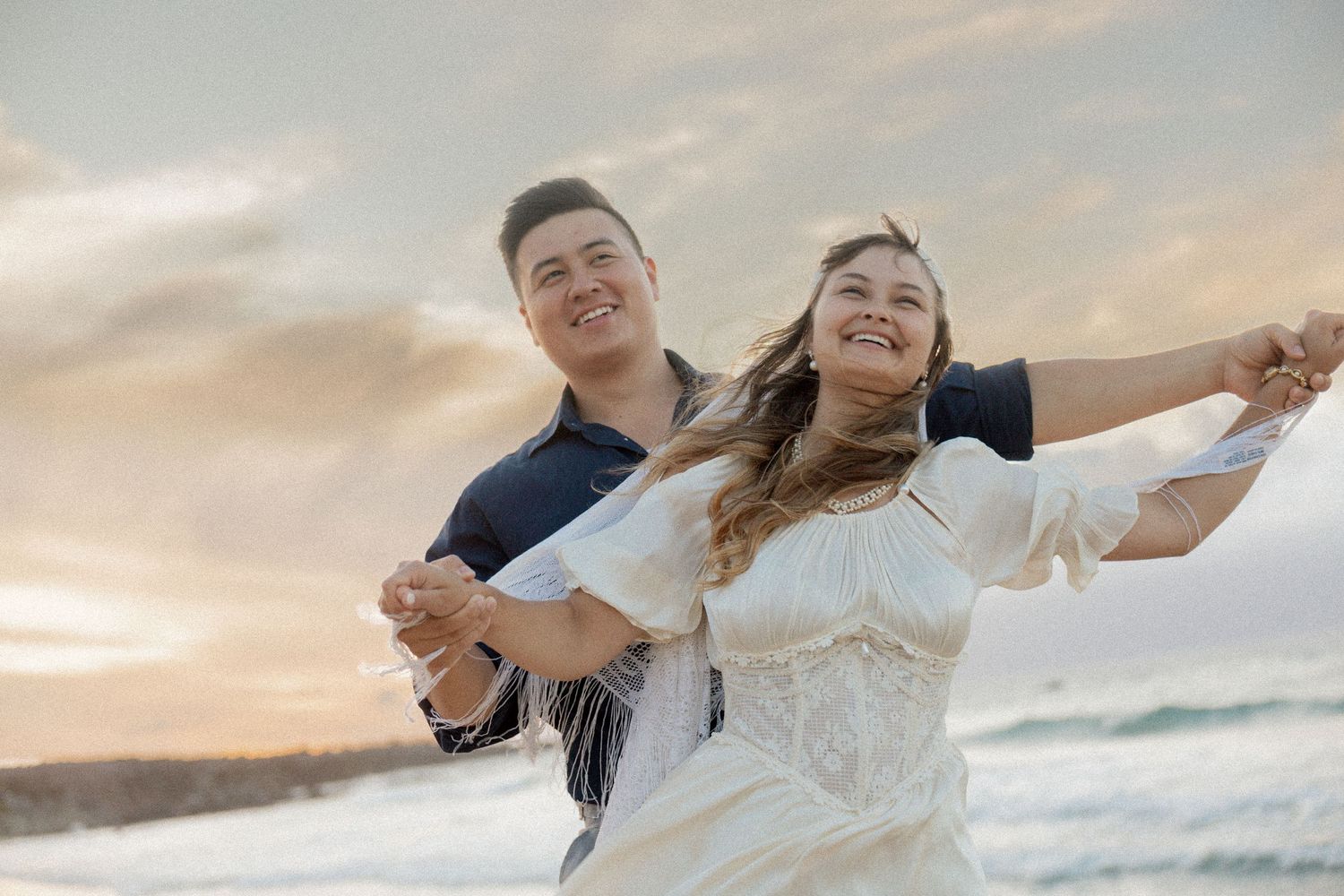 Happy couple dancing on beach at sunset, woman in white dress with arms outstretched, joyful romantic moment.