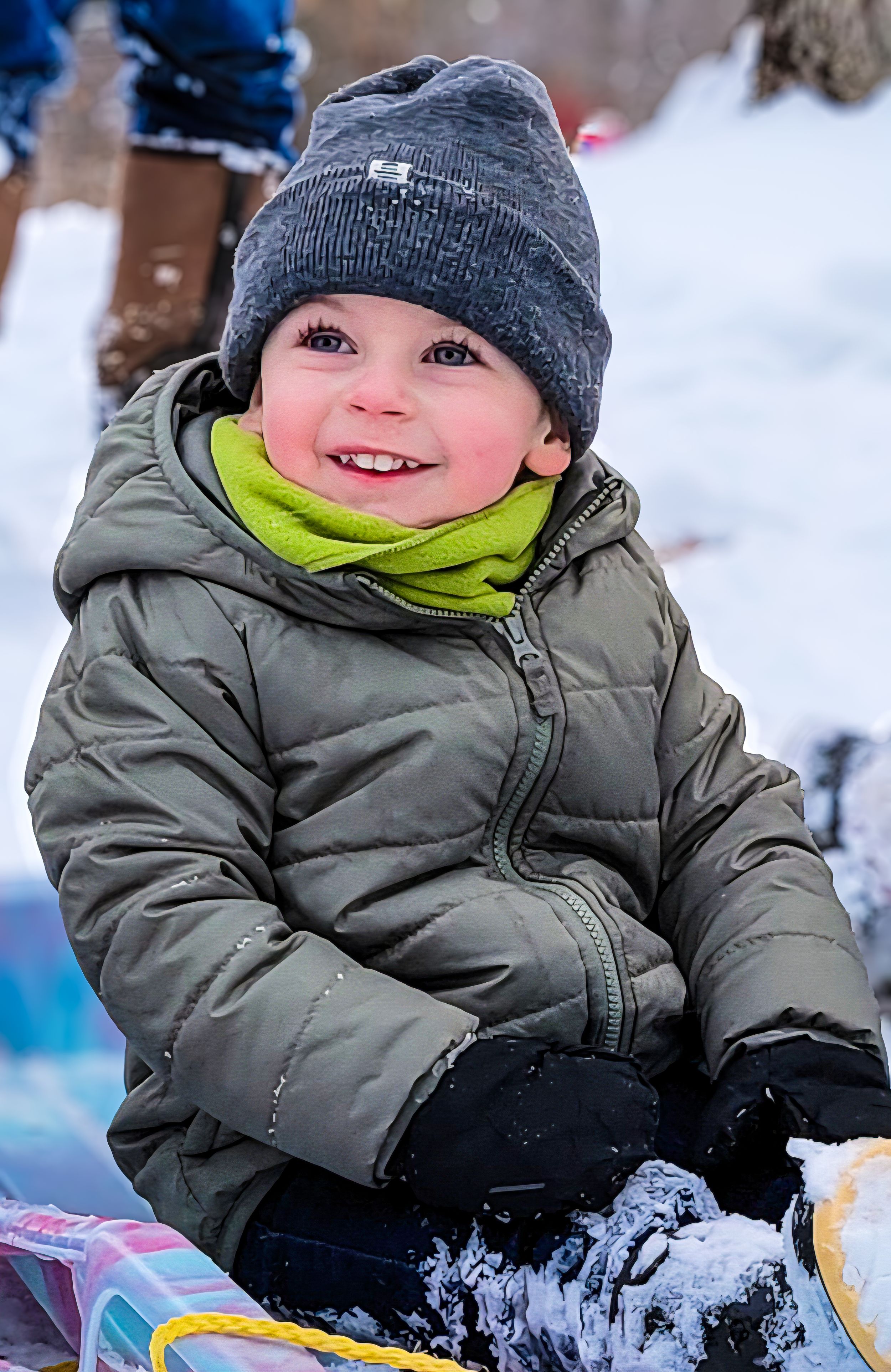 Happy toddler in gray puffer jacket and beanie smiles while playing in the snow on a winter day.