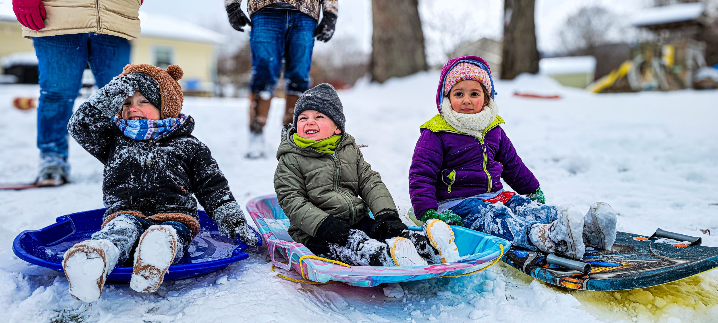 Three young children in winter gear sitting on colorful sleds in the snow, enjoying a winter sledding day outdoors.