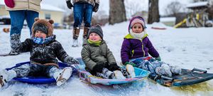 Three young children in winter gear smile while sledding in the snow on a cold winter day.