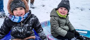 Two young children laughing and sledding in the snow, dressed in winter coats and hats on a snowy day.