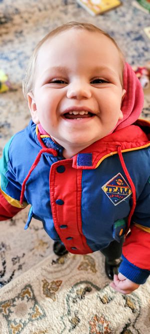 Smiling baby wearing a colorful blue and red Team jacket with a hood, looking up at the camera indoors.