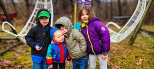 Four young children in colorful winter jackets playing together at an outdoor playground on a fall day.