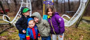 Group of four kids in bright winter coats laughing and hugging at an outdoor playground in autumn.