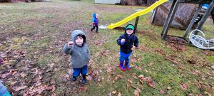 Two young children in rain boots and hooded jackets playing outdoors near a yellow slide on a cloudy fall day.