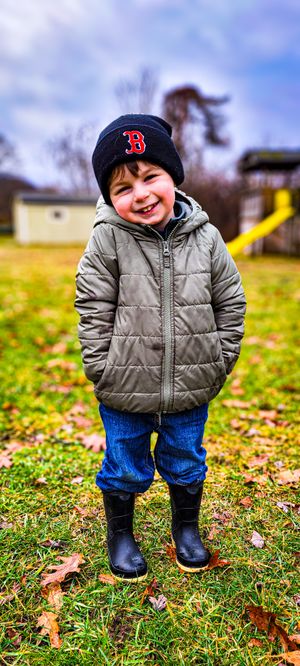 Smiling toddler boy in Boston Red Sox beanie, puffer jacket, jeans, and rain boots standing on grass with fall leaves.