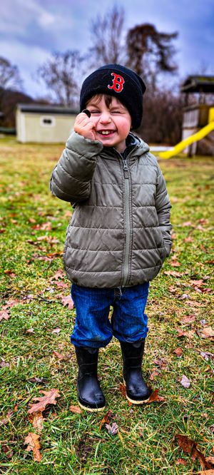 Smiling toddler in Boston Red Sox beanie, olive puffer jacket, jeans, and black rain boots standing on grass with fallen leaves.