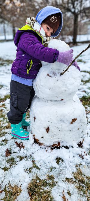 Young child in purple jacket and teal boots hugging a small snowman in a snowy yard.