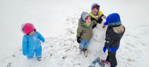 Four children in colorful winter coats building a small snowman together in a snowy outdoor setting.