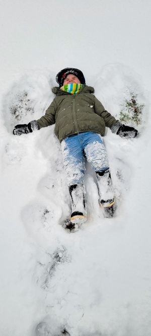 Child in green jacket and jeans making a snow angel in fresh white snow wearing colorful scarf and black hat.