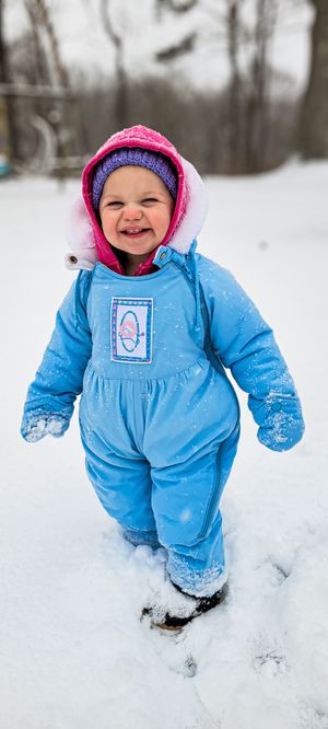 Smiling toddler girl in blue snowsuit and pink hood playing in snow during winter outdoors.