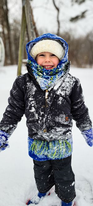 A happy young child covered in snow wearing a blue hooded jacket, scarf, and mittens, smiling outdoors in winter.