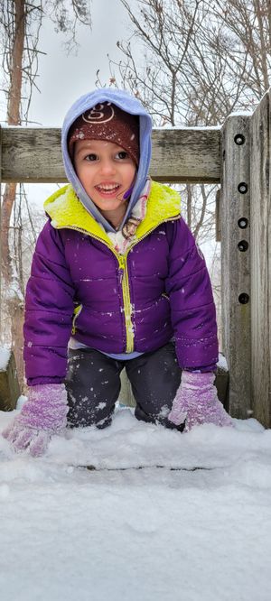 A smiling young girl in a purple puffer jacket and hood plays in the snow at a wooden playground structure.