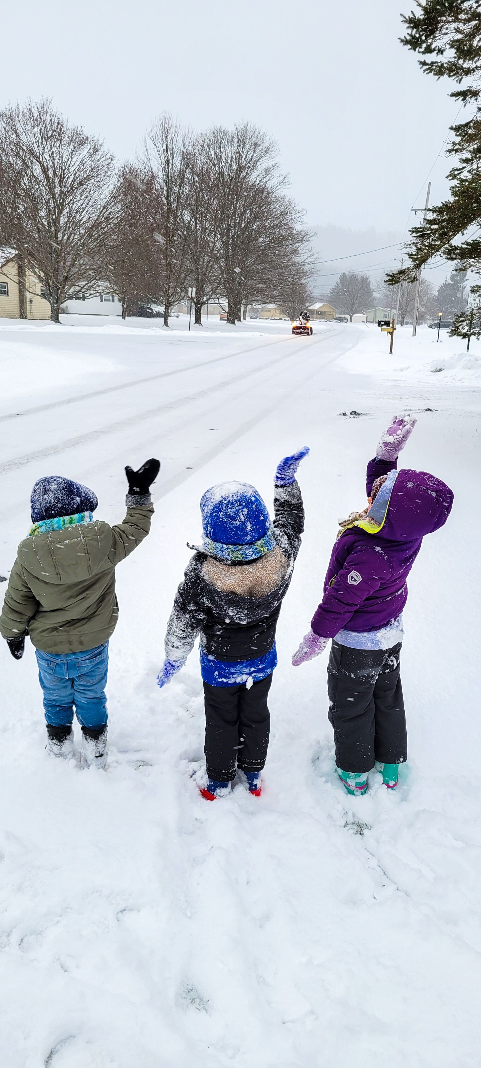 Three children in winter gear wave at a snowplow from a snowy roadside during a heavy snowfall.