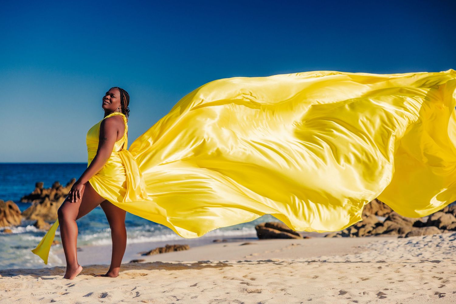 Woman in yellow flowing dress on sunny beach with ocean waves.
