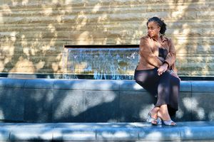 A woman in a brown jacket and black pants sits thoughtfully beside a stone fountain in sunlight.