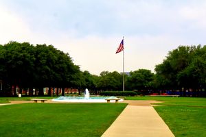 American flag flies over fountain in lush green park with tree-lined pathway on cloudy day.