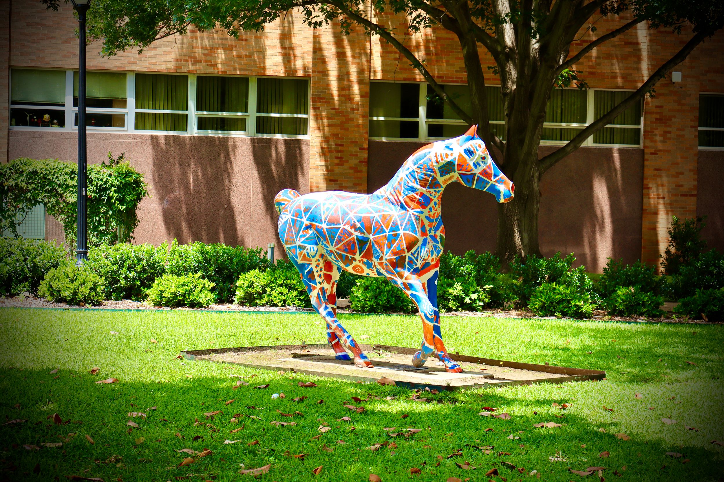 Colorful mosaic horse sculpture displayed on green lawn outside brick building on sunny day.