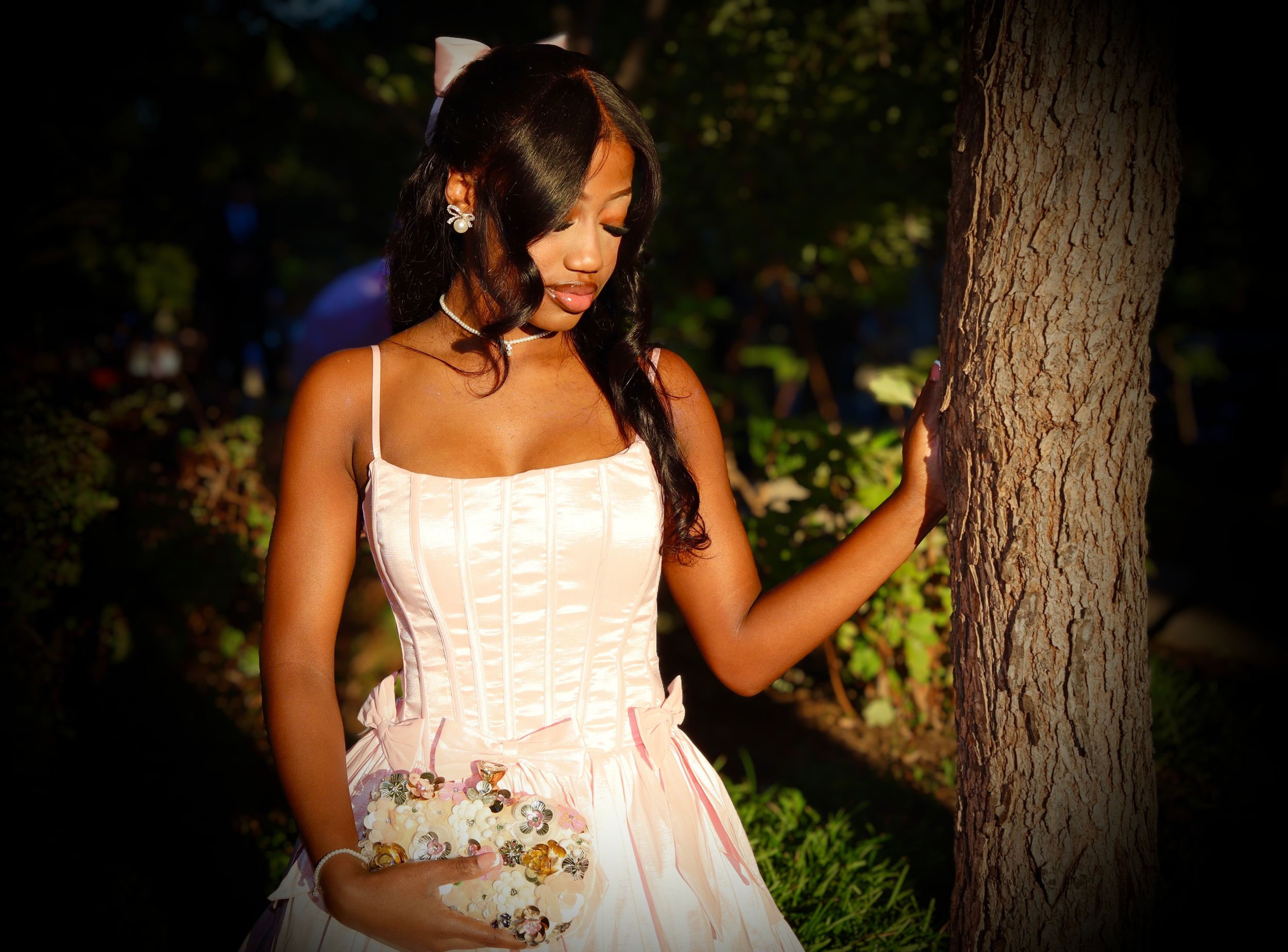 Young woman in a pink corset-style gown with a floral bouquet leans against a tree trunk in a softly lit outdoor garden at dusk.