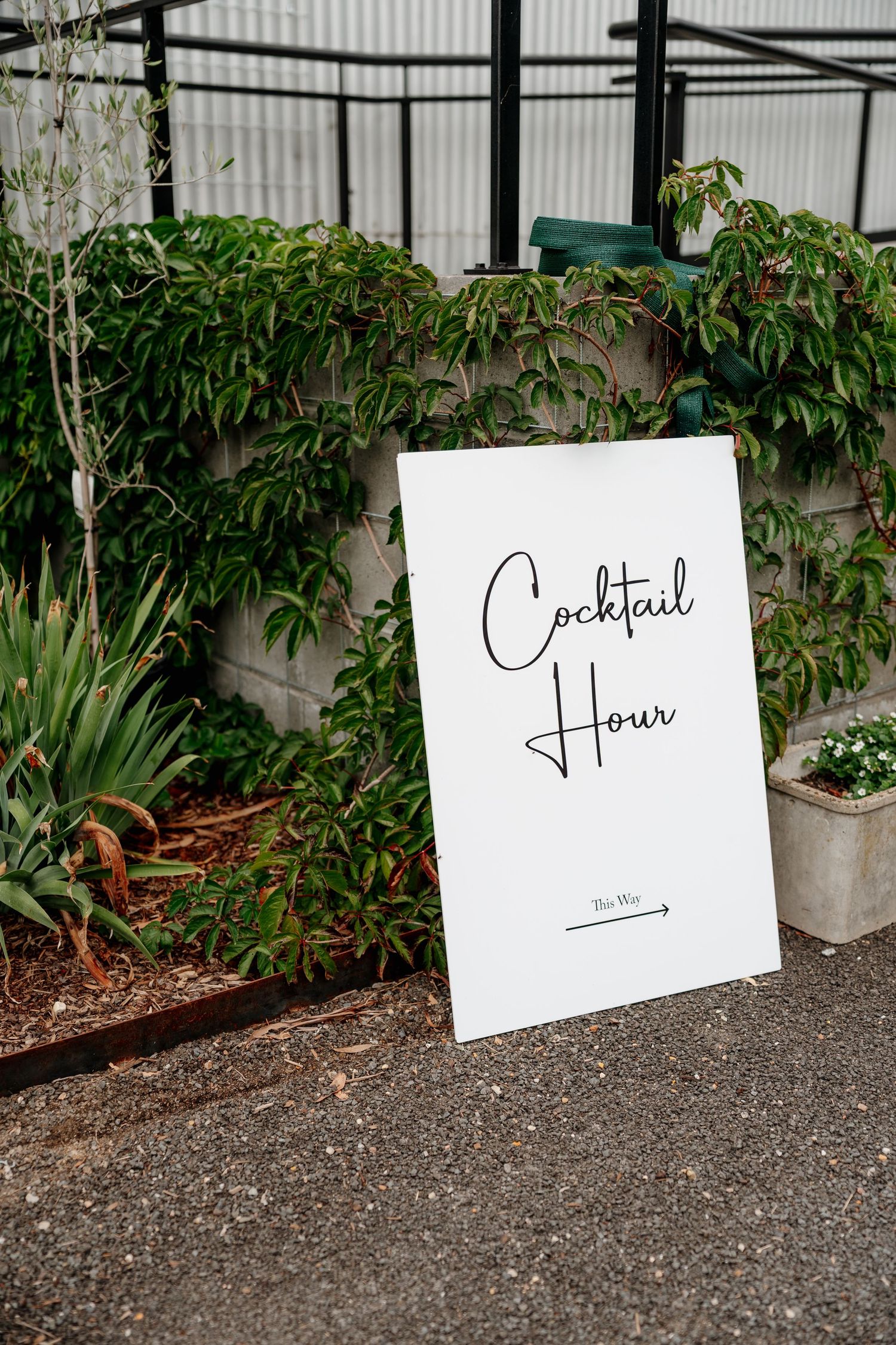 White cocktail hour wedding sign leaning against a lush green garden wall on a paved outdoor venue.