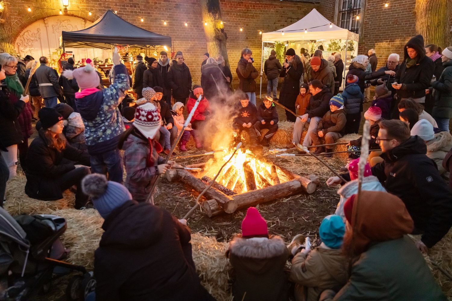 Crowd gathered around a large bonfire at an outdoor winter market, with market stalls and string lights in background.