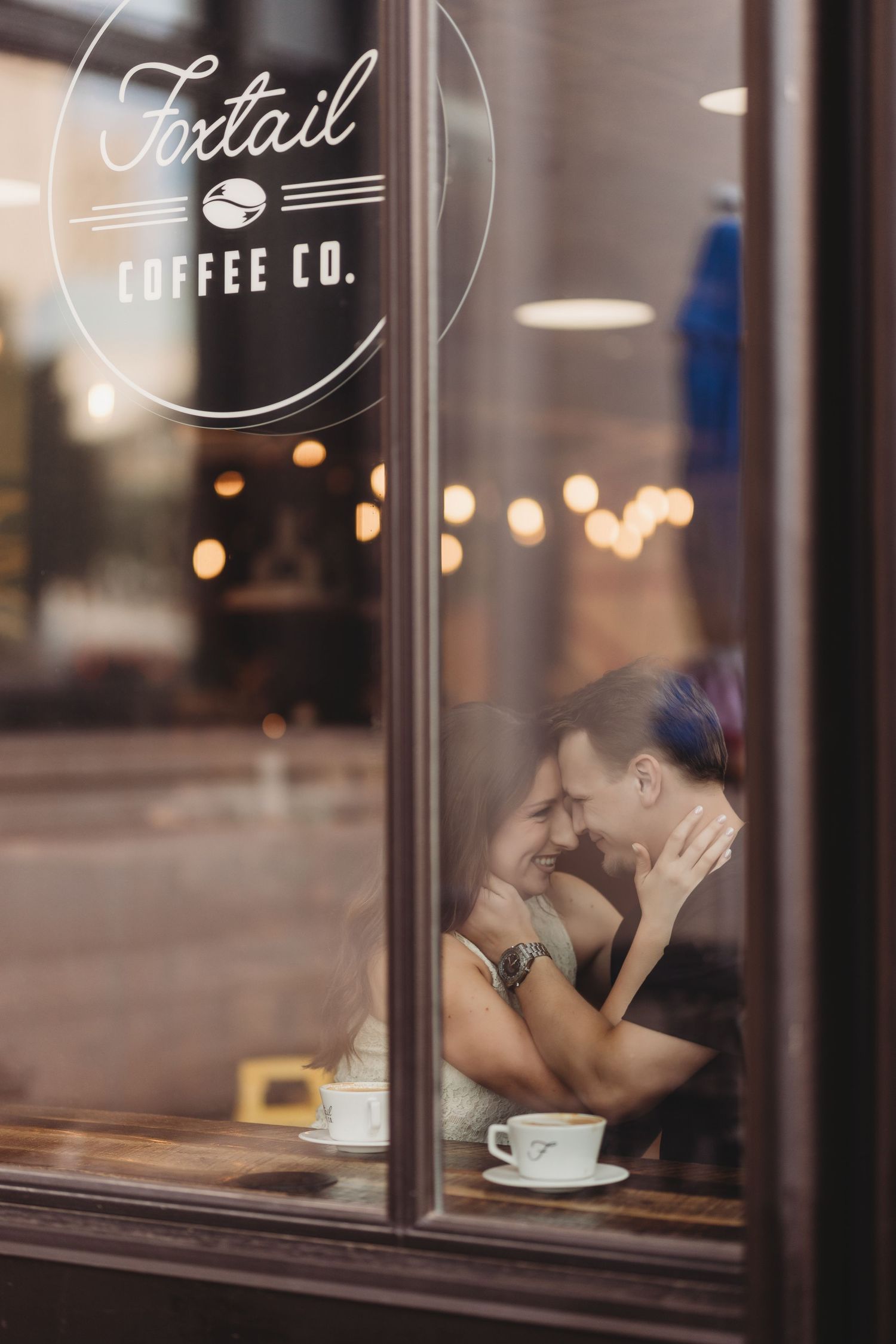 Couple sharing romantic moment at Foxtail Coffee Co., seen through window with coffee cups on table.