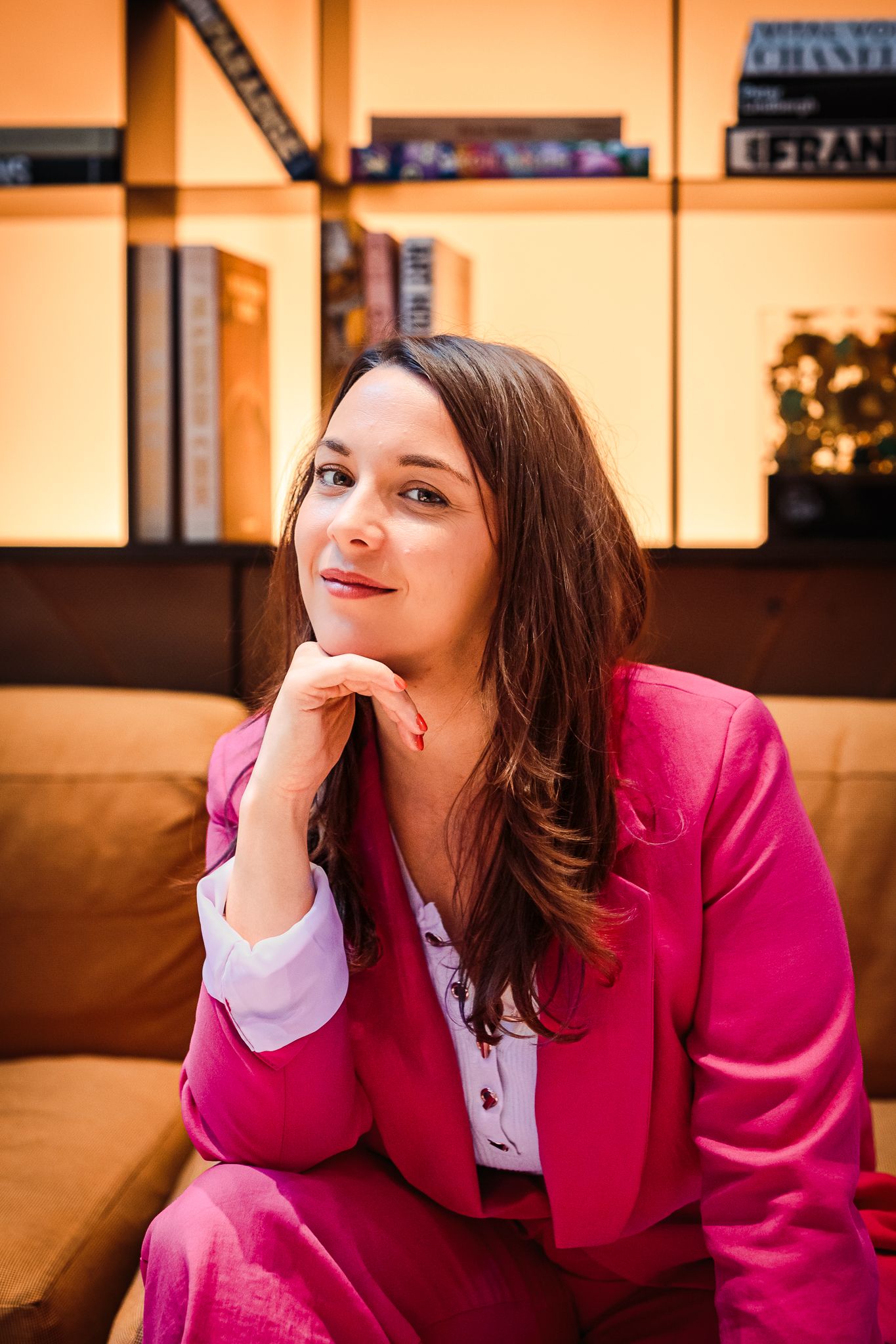 Smiling woman in red satin bomber jacket and gold earrings sitting in velvet chair.