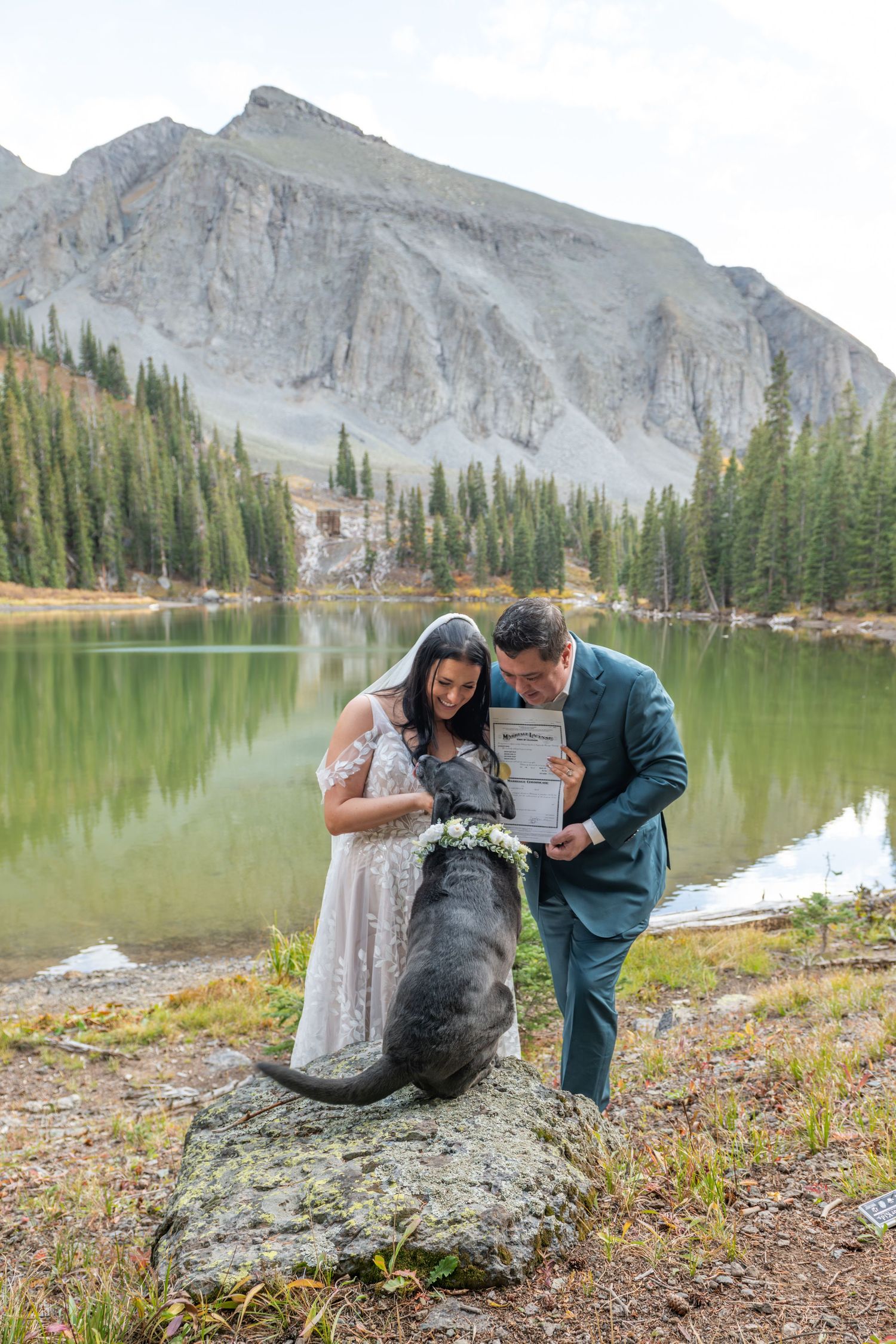 A bride and groom show their dog their marriage license by a mountain lake, surrounded by pine trees and rocky peaks.