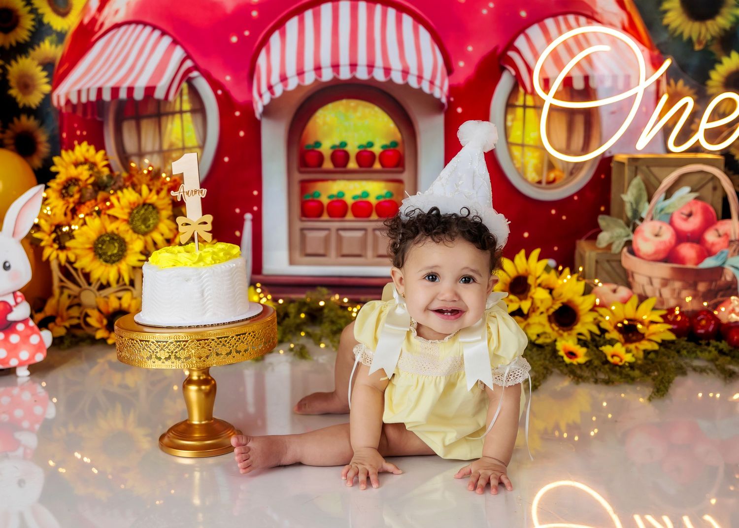Baby girl smiling at her first birthday cake smash with sunflowers and a fairytale house backdrop.