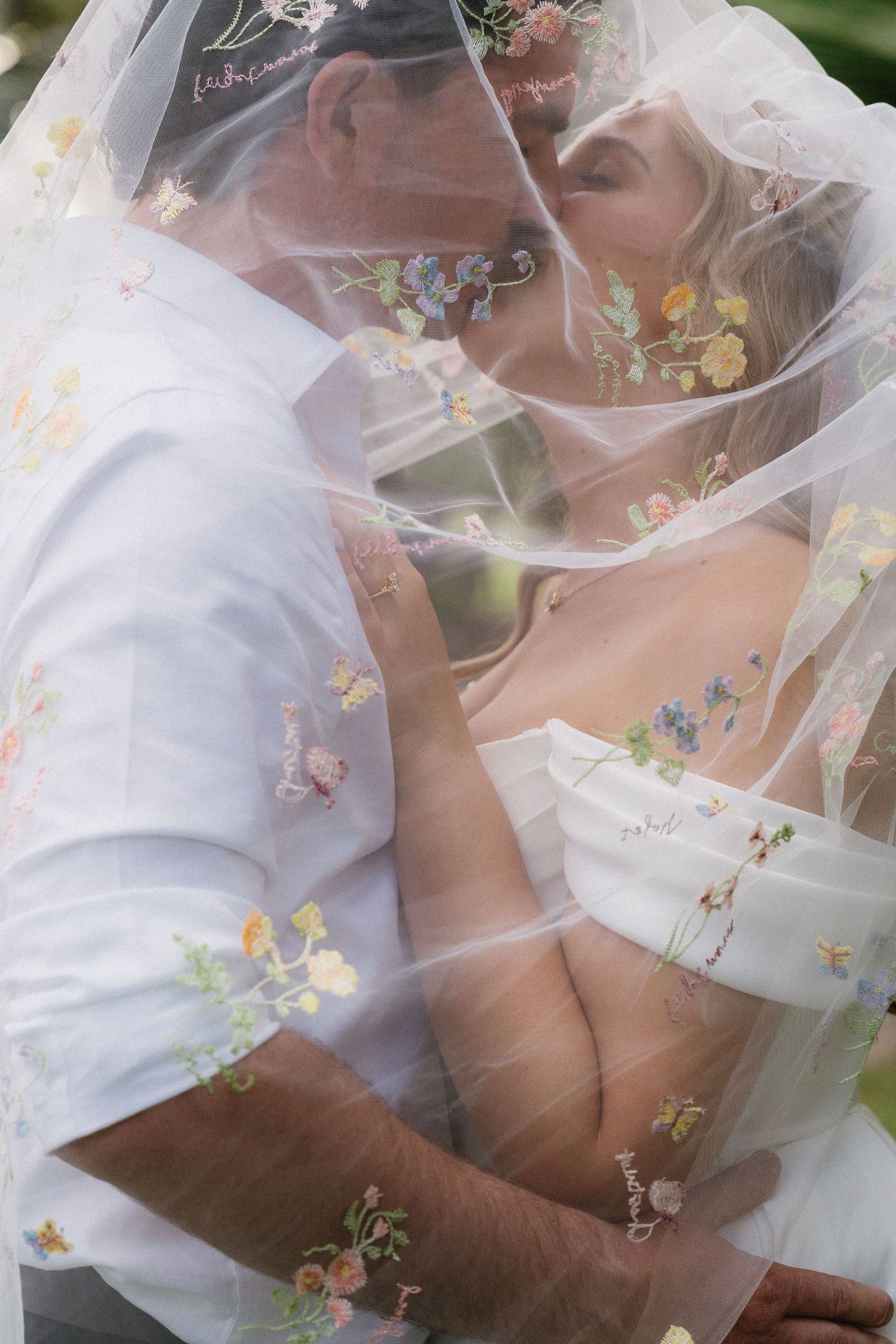 Bride and groom kissing under a floral embroidered wedding veil, captured in a romantic close-up portrait.
