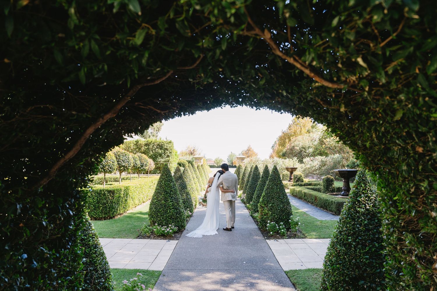 Couple walking hand-in-hand through an elegant garden archway lined with sculpted topiaries on their wedding day.