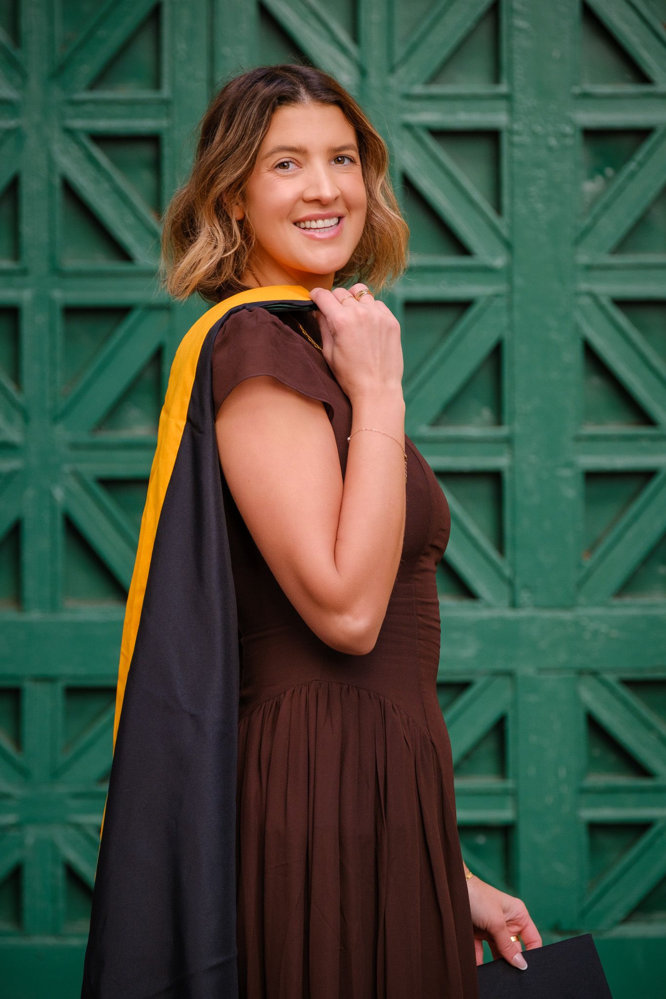 Smiling graduate woman in brown dress and black and gold academic regalia poses outdoors by green lattice wall.