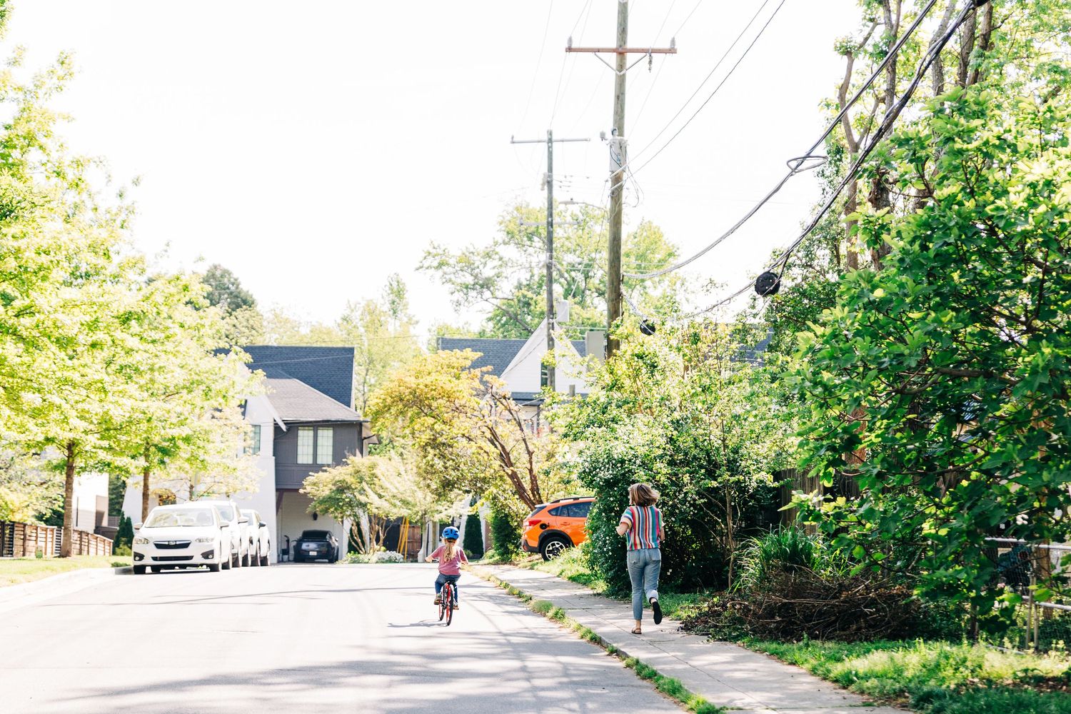 A mother jogging to keep up with her child on a bike on a quiet tree-lined residential street on a sunny day.