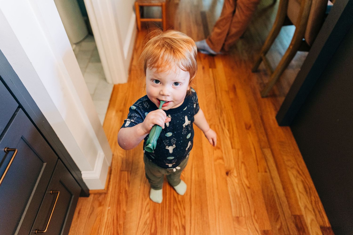 Adorable toddler with red hair standing on hardwood floor, looking up at camera while brushing his teeth.