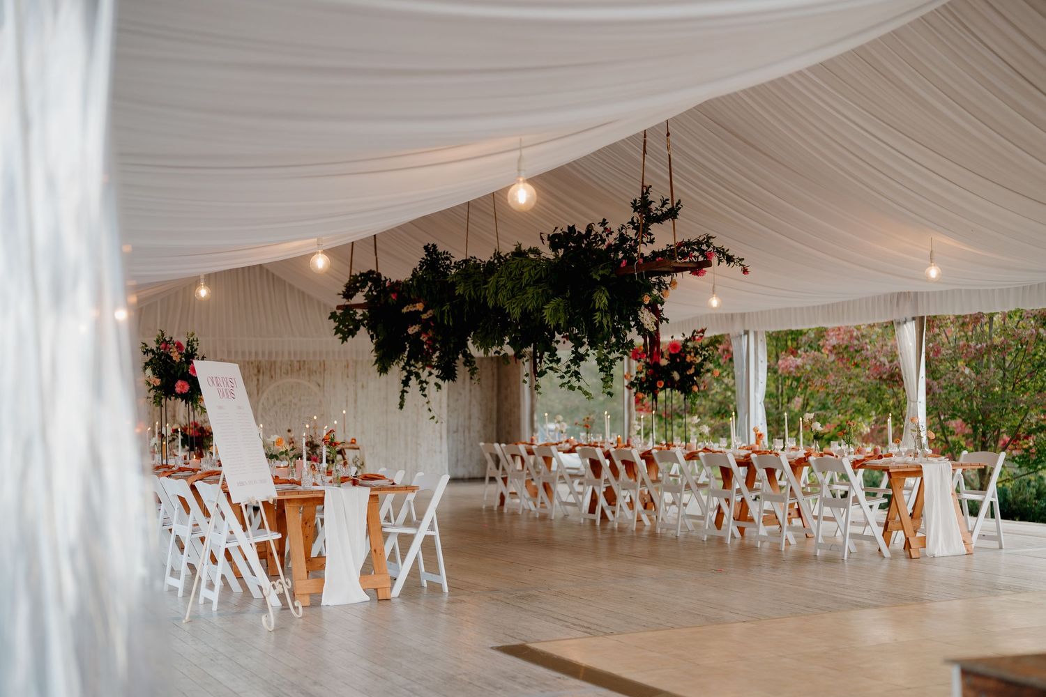 Elegant outdoor wedding reception tent with white draping, string lights, lush floral chandeliers, and white chairs at rustic wood tables.
