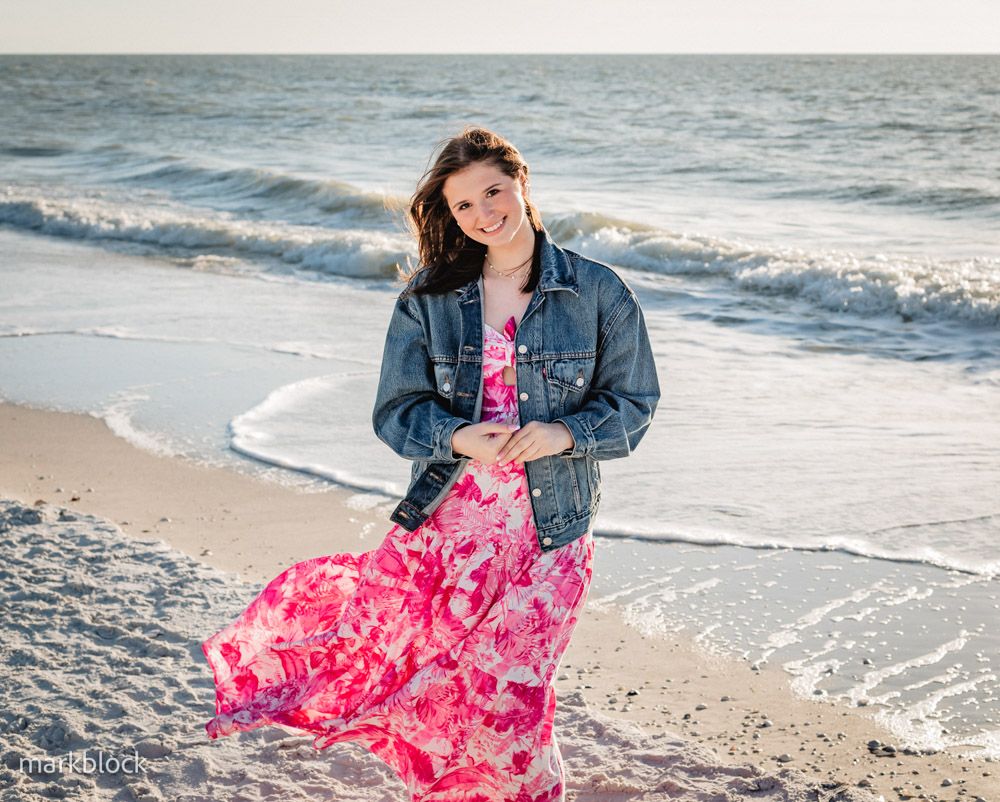 Young woman in pink floral maxi dress and denim jacket smiling on a sandy beach with ocean waves behind her.