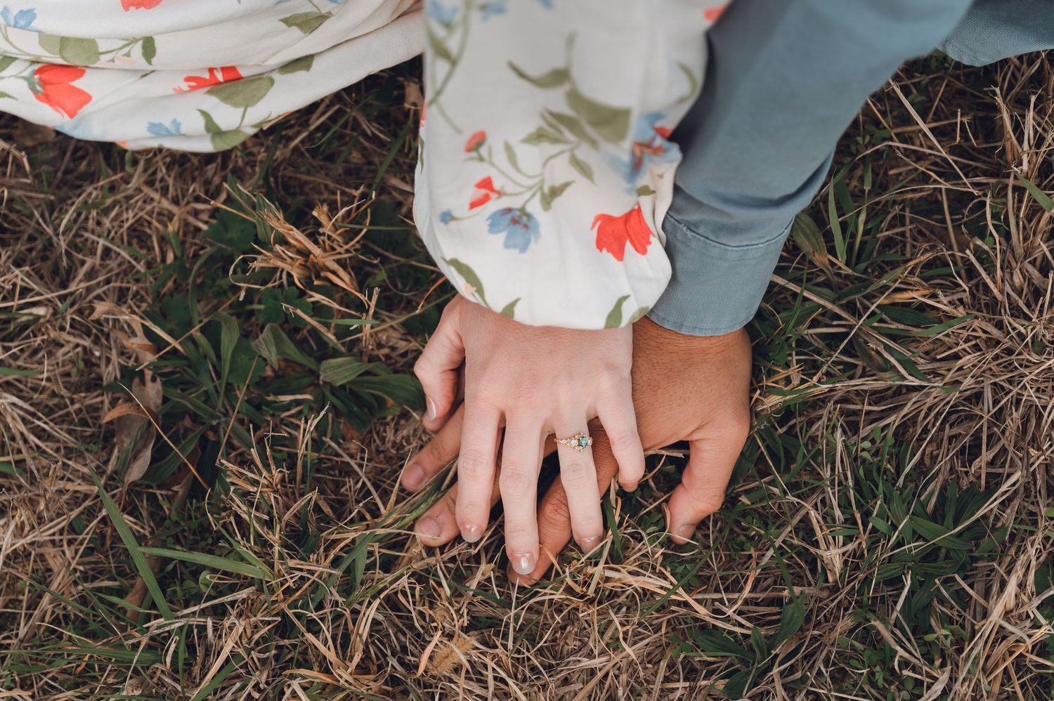 Two hands resting on grass, one wearing a ring, with a floral dress sleeve visible in a romantic outdoor setting.