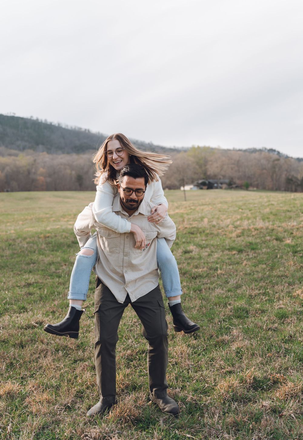 A cheerful woman rides piggyback on a bearded man wearing glasses in a green field with hills in the background.