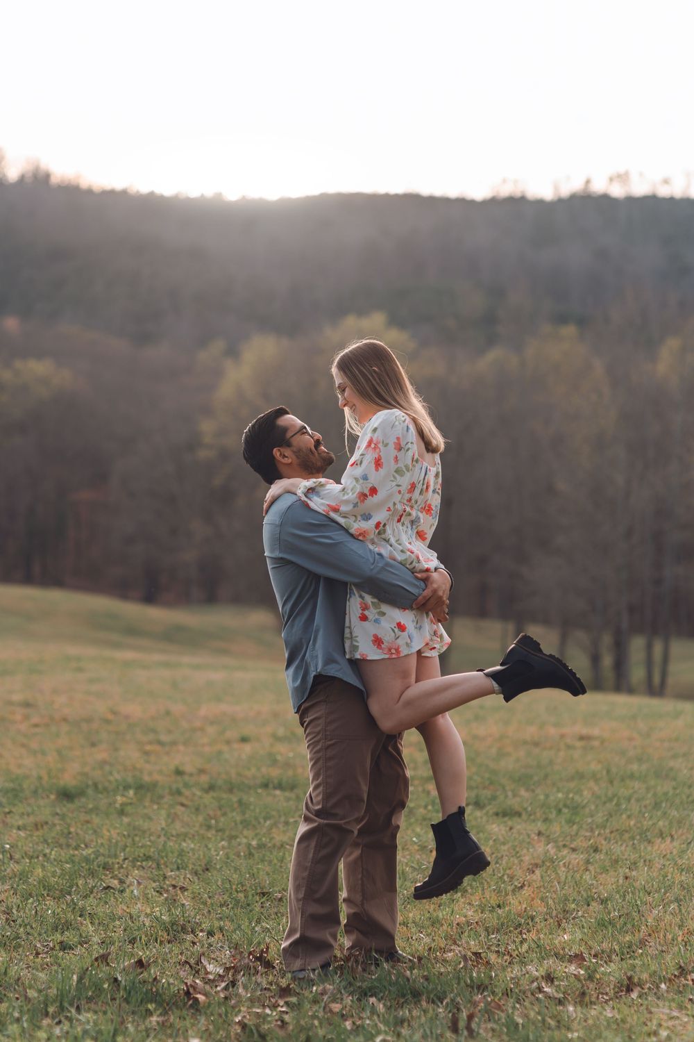 A man lifts a woman in a floral dress outdoors on a grassy field with autumn trees and sunset in the background.
