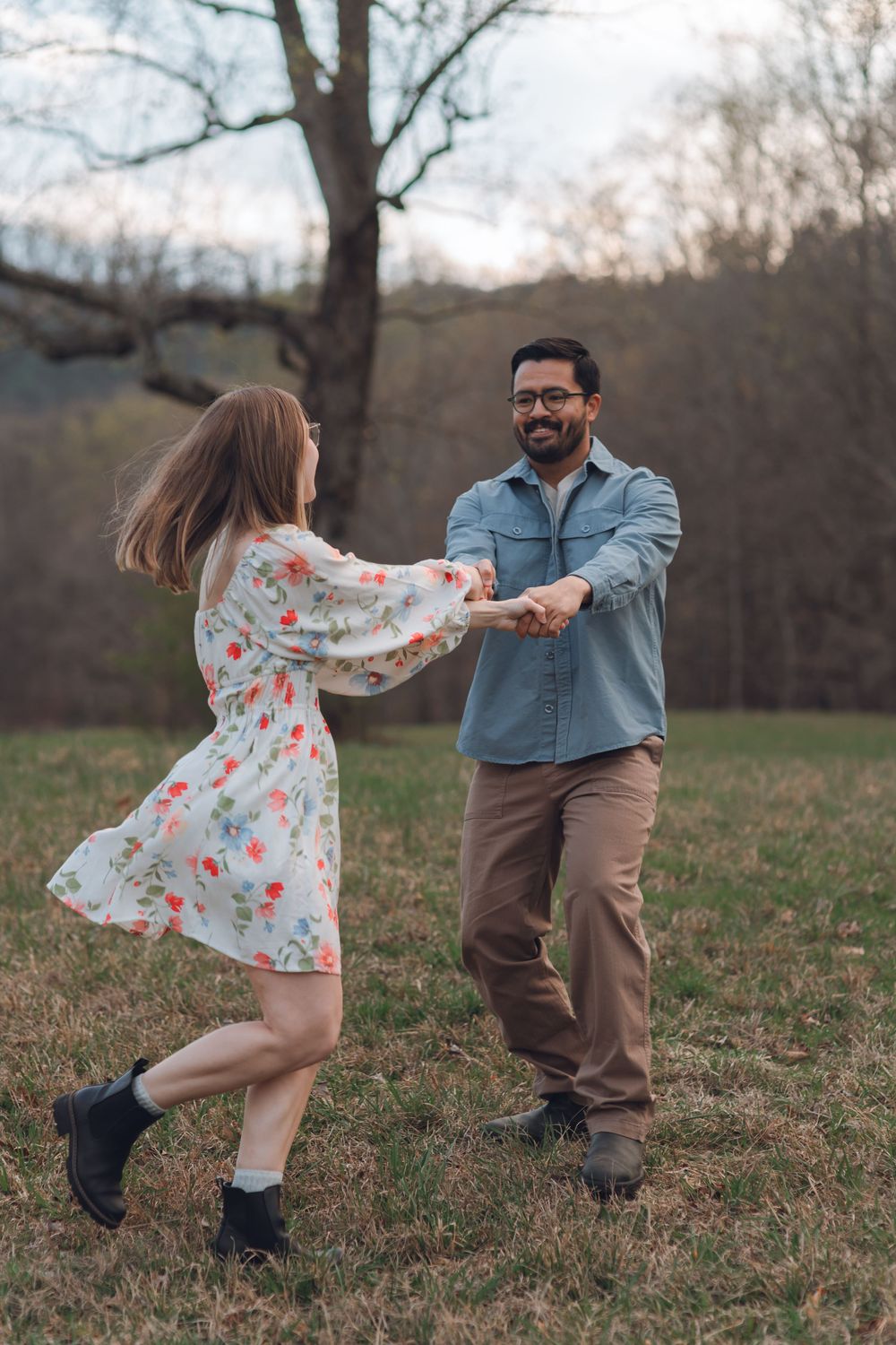 A joyful couple dances together in a grassy field, the woman in a floral dress and the man in a denim shirt.