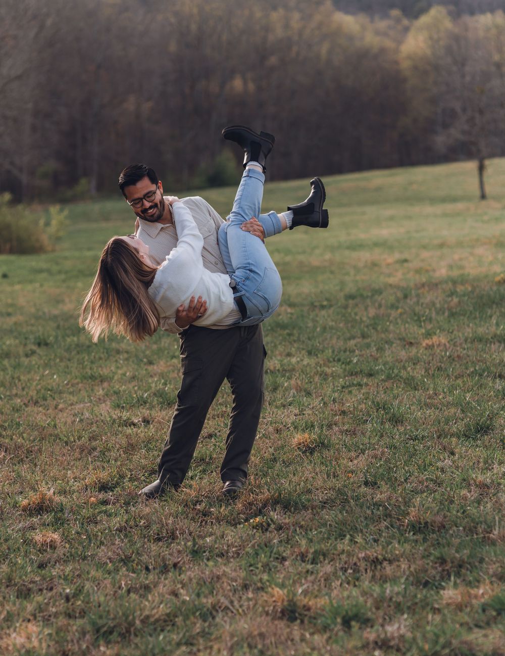 A man joyfully lifts a woman in a grassy field, both smiling during a romantic outdoor couples photo session.