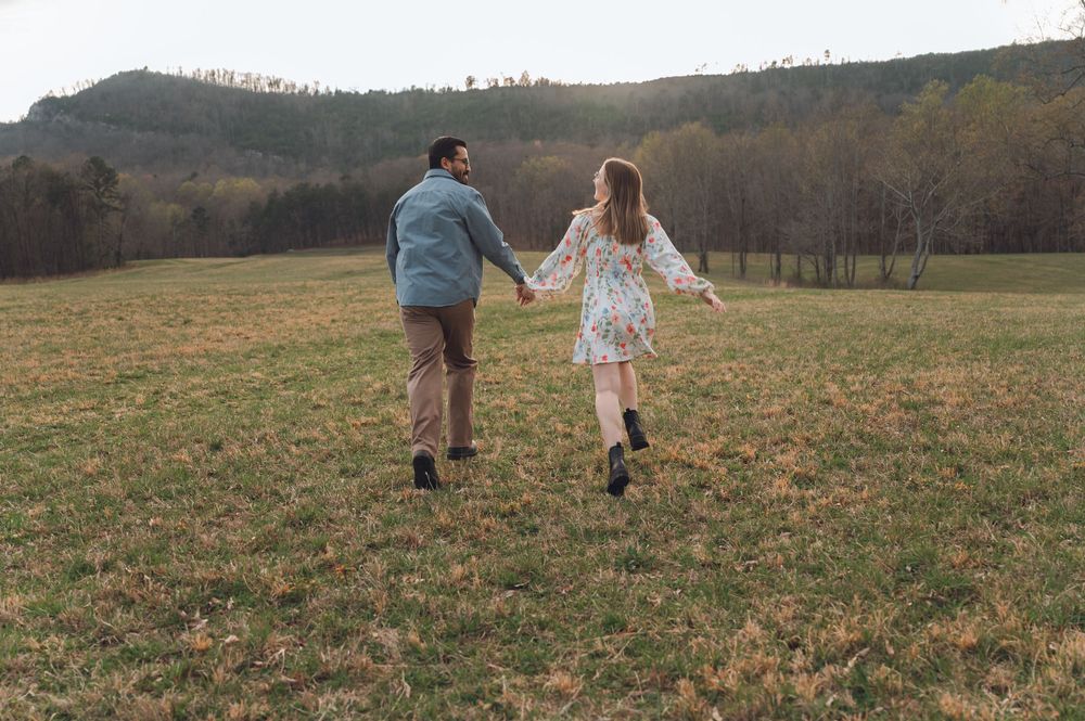 Couple holding hands running through a green meadow with rolling hills in the background.