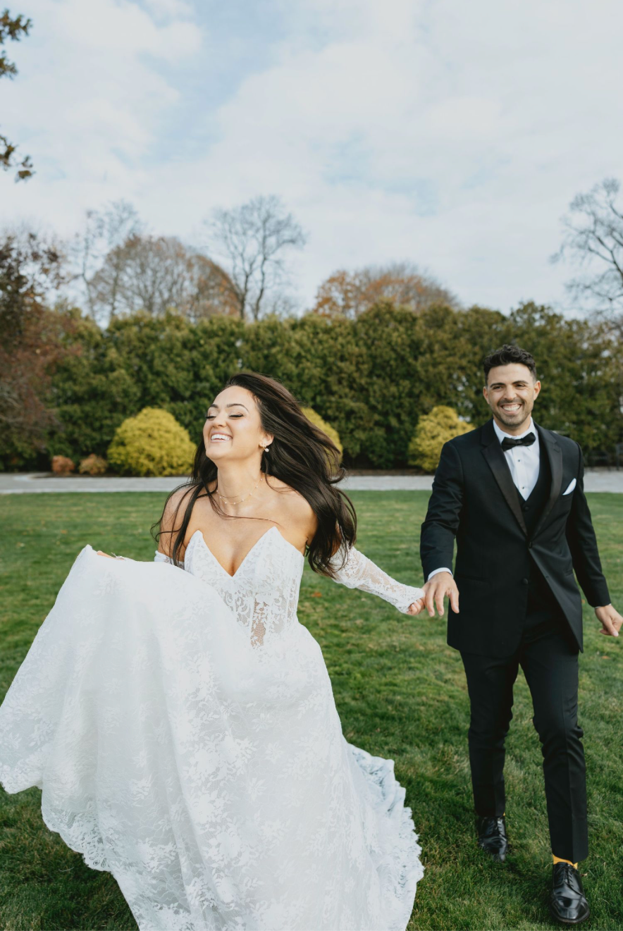 A joyful bride in a lace gown holds hands with her groom in a black tuxedo as they run across a green lawn.