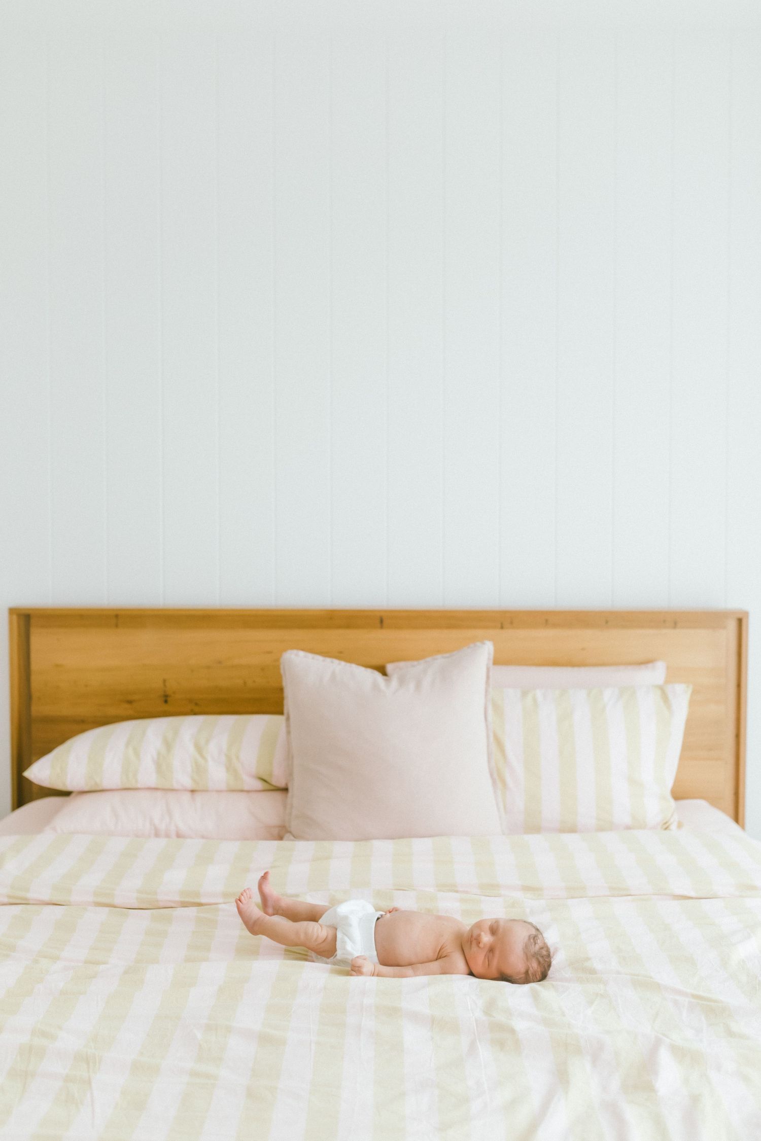 Newborn baby in diaper lying on striped bedding against wooden headboard with pink pillows.