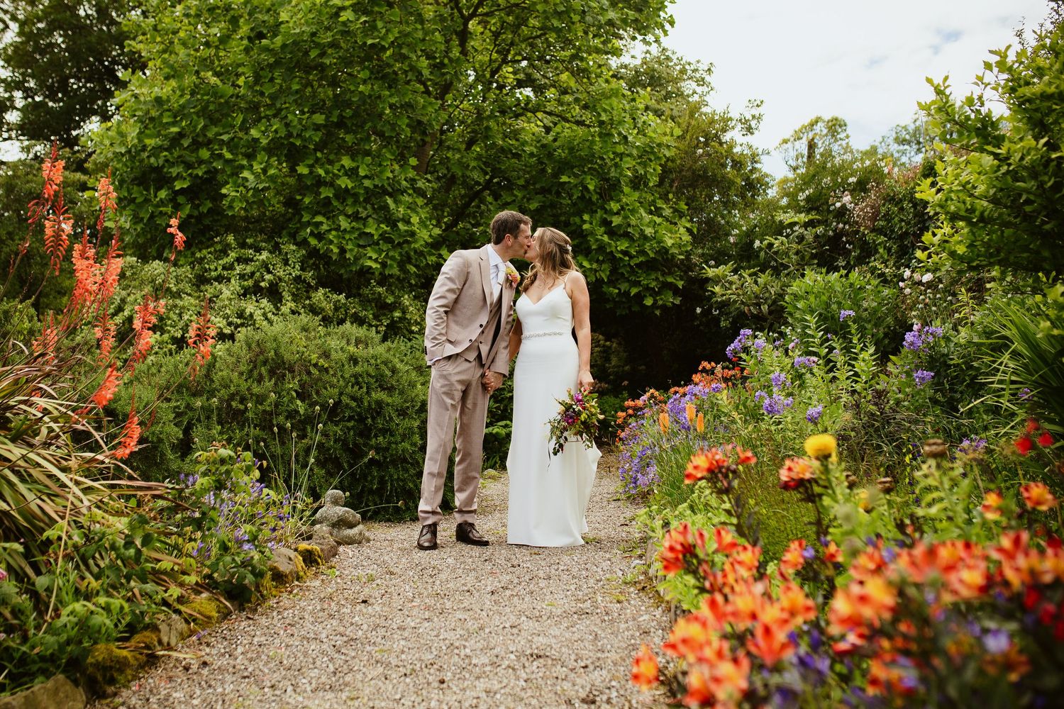 Bride and groom share a romantic kiss on a garden path surrounded by vibrant colorful flowers and lush greenery.