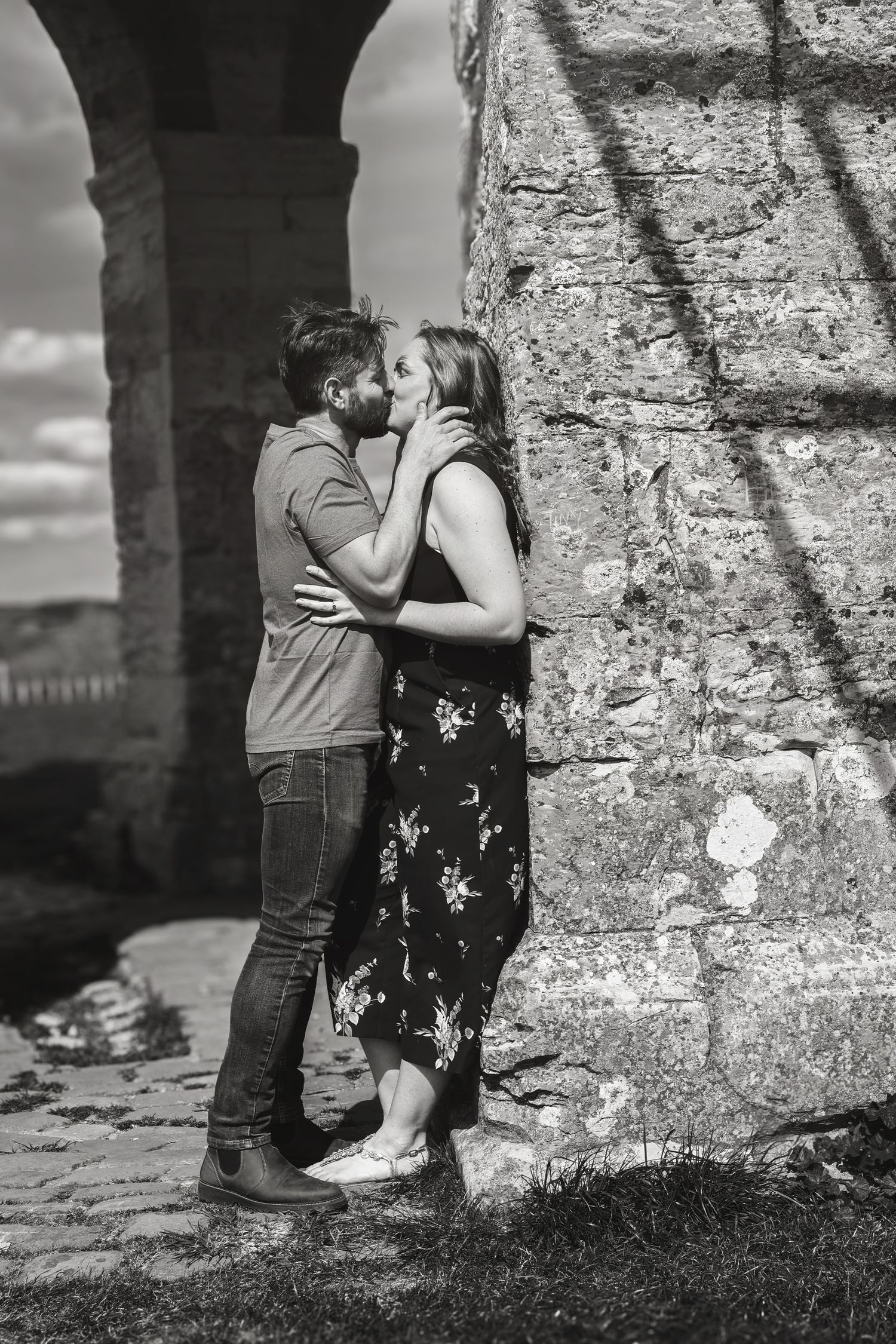 Black and white photo of a romantic couple kissing against ancient stone ruins outdoors.