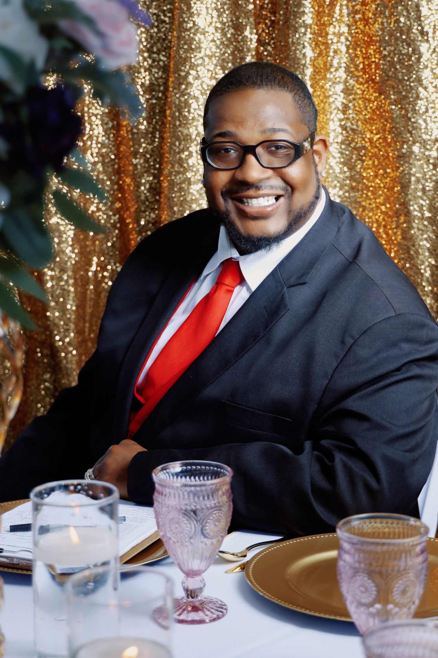 Smiling Black man in a black suit and red tie sits at an elegant table with gold sequin backdrop.
